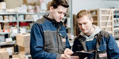 In einem Lagerraum stehen zwei Männer, die konzentriert auf ein Tablet schauen, umgeben von Kisten und Regalen.