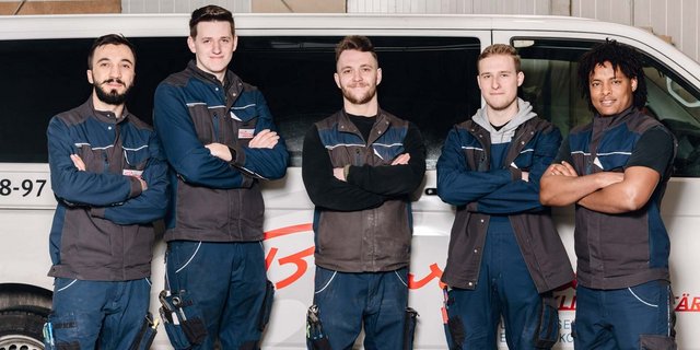 Fünf junge Männer in blauen Arbeitsanzügen stehen stolz vor einem Fahrzeug, das für ihre Arbeit steht, und strahlen Zuversicht aus.