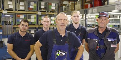 In einer gut organisierten Werkstatt stehen fünf Männer, die freundlich in die Kamera schauen, während sie ihre Arbeitskleidung tragen.