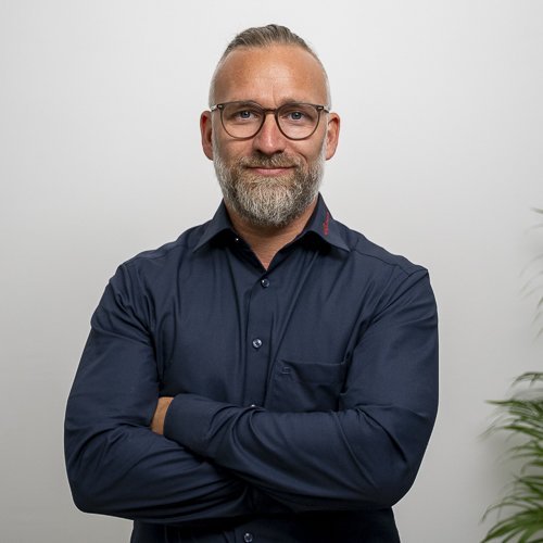 In einem modernen Raum posiert ein Mann mit Brille, der eine freundliche Ausstrahlung hat und seine Arme verschränkt.