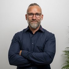 Professionelles Porträt eines lächelnden Mannes mit Bart In einem modernen Raum posiert ein Mann mit Brille, der eine freundliche Ausstrahlung hat und seine Arme verschränkt.