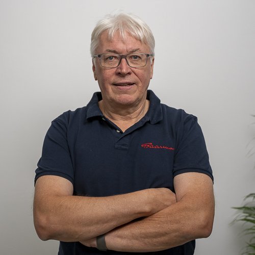 Ein älterer Mann mit Brille und grauen Haaren steht mit verschränkten Armen vor einer neutralen Wand.