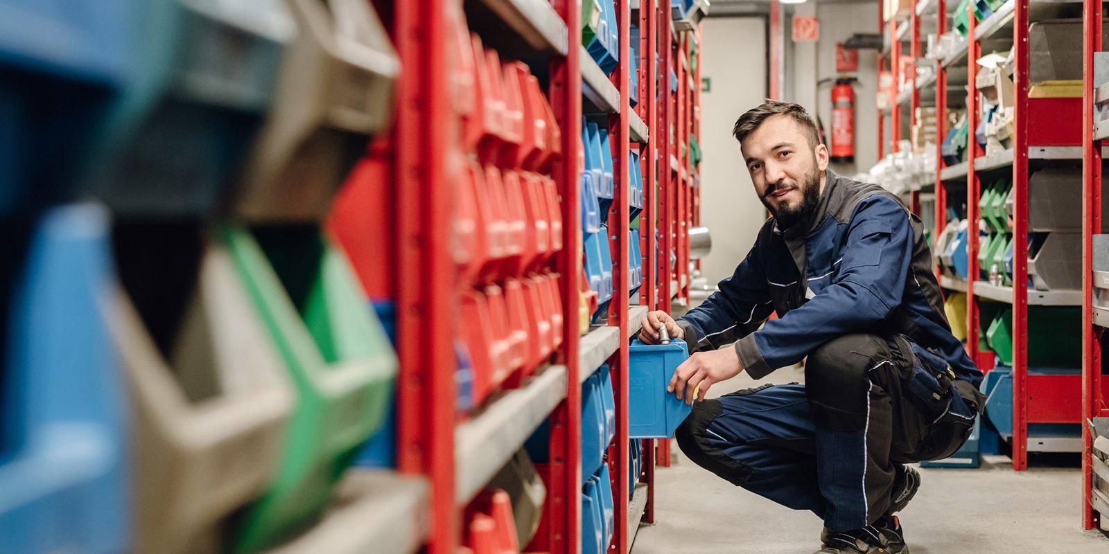 In einem gut organisierten Lagerraum sortiert ein Arbeiter blaue Kisten und zeigt dabei ein freundliches Lächeln.