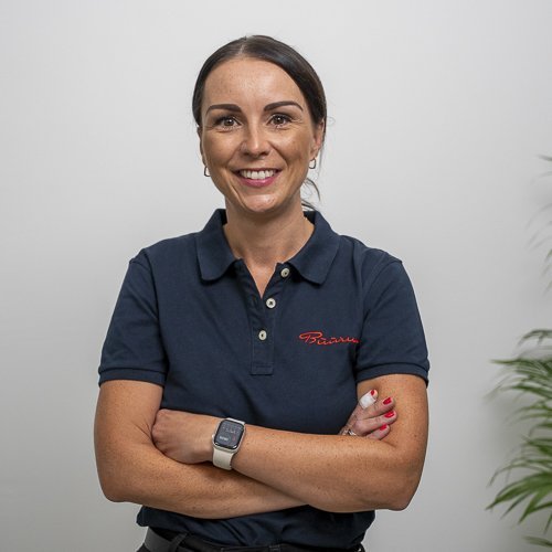 In einem hellen Raum posiert eine Frau mit einem selbstbewussten Ausdruck, die ihre Arme vor dem Körper verschränkt.