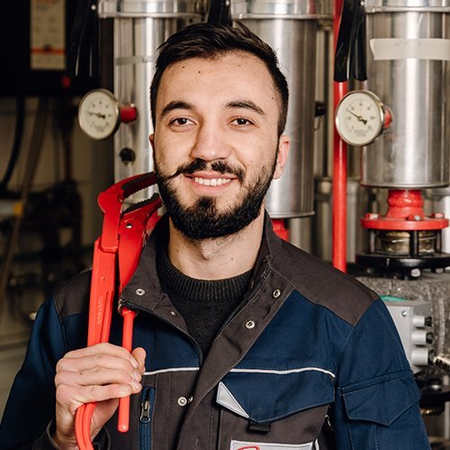 Ein junger Handwerker mit Bart lächelt, während er eine Rohrzange in einer Werkstatt hält, umgeben von Maschinen.