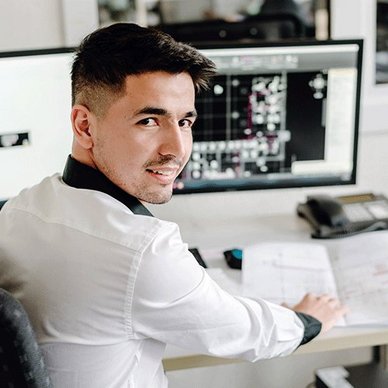 In einem modernen Büro lächelt ein Mann, während er an einem Computer arbeitet und technische Dokumente durchblättert.