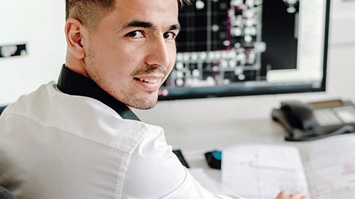 In einem modernen Büro lächelt ein Mann, während er an einem Computer arbeitet und technische Dokumente durchblättert.