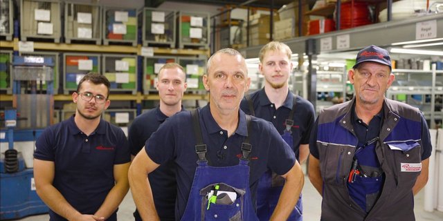 Fünf Mitarbeiter von Büürma stehen in Arbeitskleidung in einer Werkstatt vor Regalen mit Material