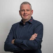 Selbstbewusster Mann mit verschränkten Armen vor neutralem Hintergrund Der Mann trägt ein dunkelblaues Hemd und hat einen selbstbewussten Ausdruck, während er in die Kamera schaut.