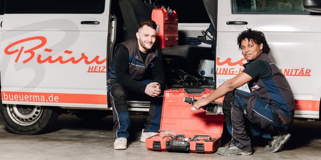 Zwei Handwerker posieren lächelnd vor einem Lieferwagen, umgeben von Werkzeugen und Ausrüstung in einer Werkstatt.