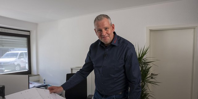 In einem hellen Büro steht ein Mann mit grauen Haaren, der freundlich in die Kamera schaut und Papiere in der Hand hält.