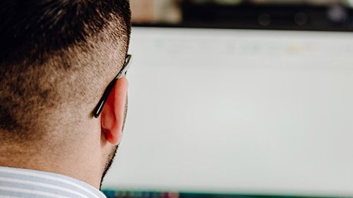 Der Rücken eines Mannes mit einem modernen Haarschnitt ist zu sehen, während er auf einen unbeschrifteten Computerbildschirm schaut.