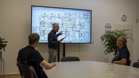 Ein Mann präsentiert technische Pläne auf einem großen Bildschirm, während zwei Zuhörer aufmerksam im Konferenzraum sitzen.