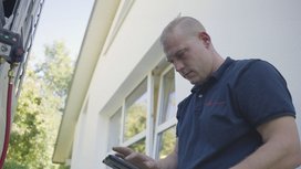 Ein Techniker überprüft mit einem Tablet die Einstellungen eines Geräts an der Außenwand eines Hauses.